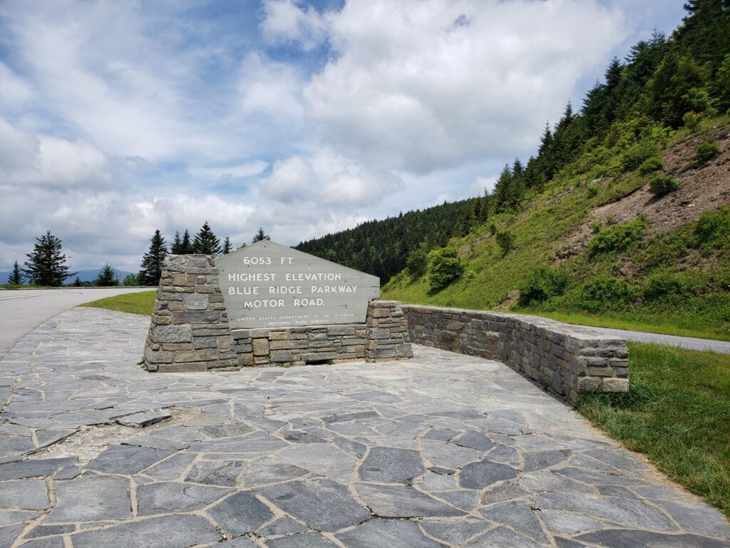 The gray sign at Richland Balsam boasts the Parkway's highest point (6,053 feet), with evergreens on the ridge to the right of the sign and a mountainous vista to the left.