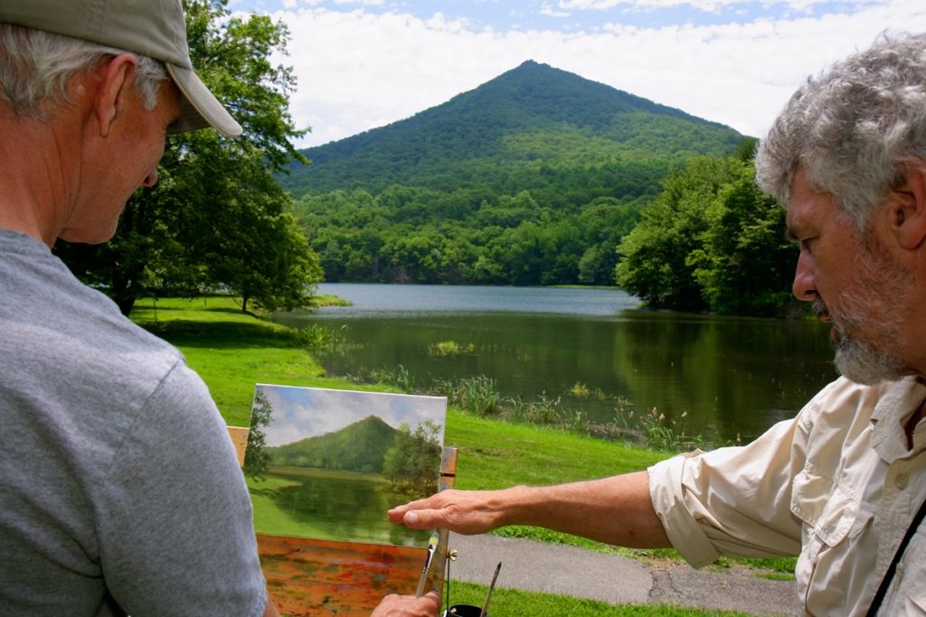 Two men sit in front of a painting of Sharp Top Mountain and Abbott Lake at Peaks of Otter, with the peak and lake in the background..