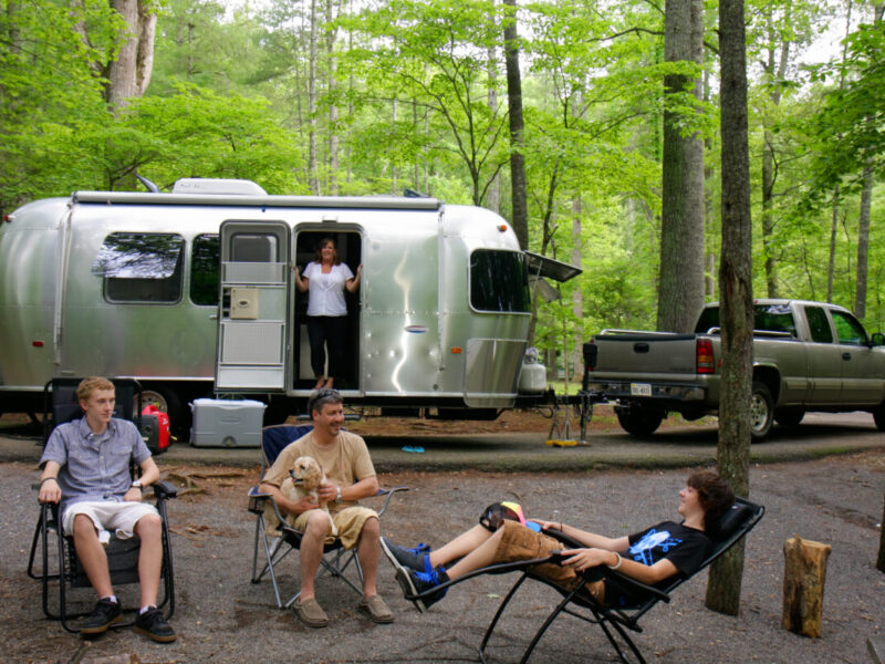 Several campers relax in chairs in front of an Airstream trailer in a shaded campsite.