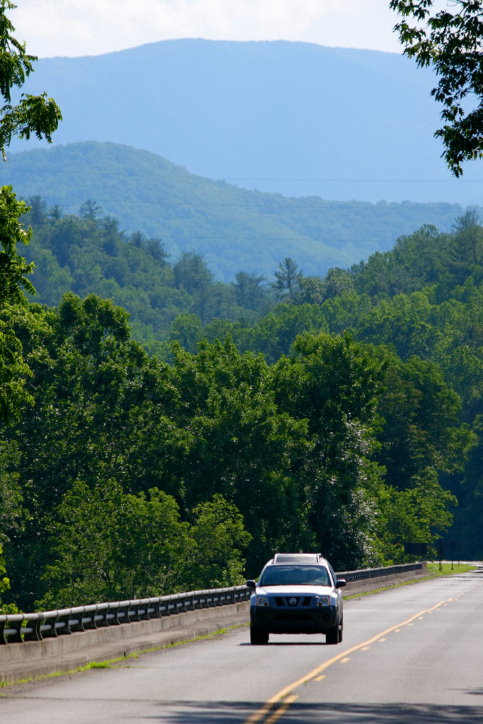 An SUV drives across the James River Bridge with Appalachian hardwoods on the riverbank and ridges beyond.