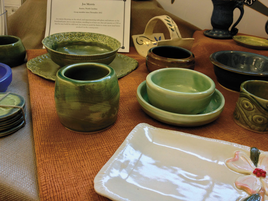 Pottery platters, bowls, coasters and other dishes are scattered on a cloth-covered tabletop with the local potter's description nearby.