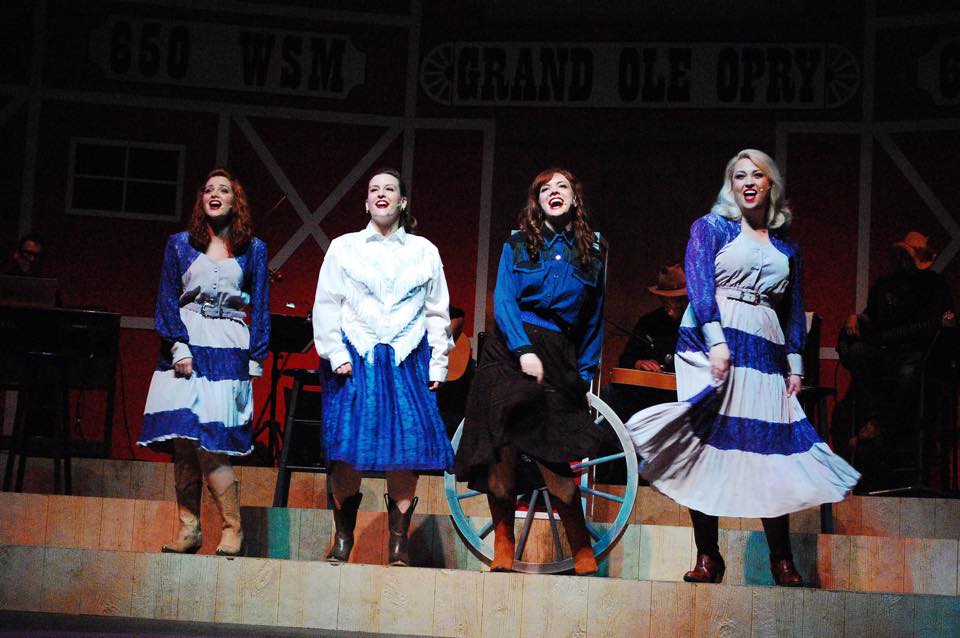 Four female singers dressed in blue and white country garb sing on stage at Wohlfahrt Haus Dinner Theatre.