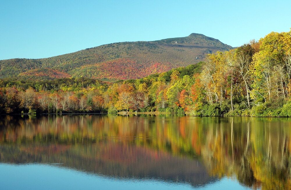 Fall-colored leaves on the trees on the shore and nearby slope reflect in the lake water below.