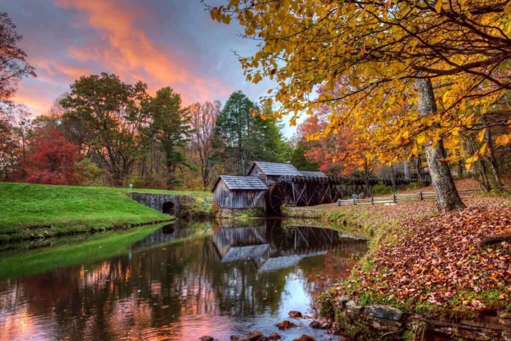 The sun sets behind the historic gristmill at Mabry Mill in Autumn, with pink highlights in the sky and red and yellow leaves framing the millpond.