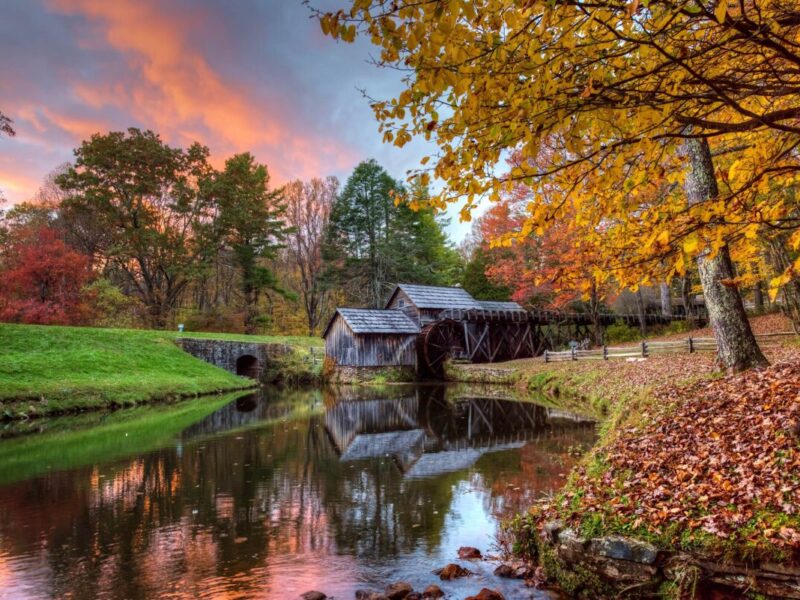 The sun sets behind the historic gristmill at Mabry Mill in Autumn, with pink highlights in the sky and red and yellow leaves framing the millpond.