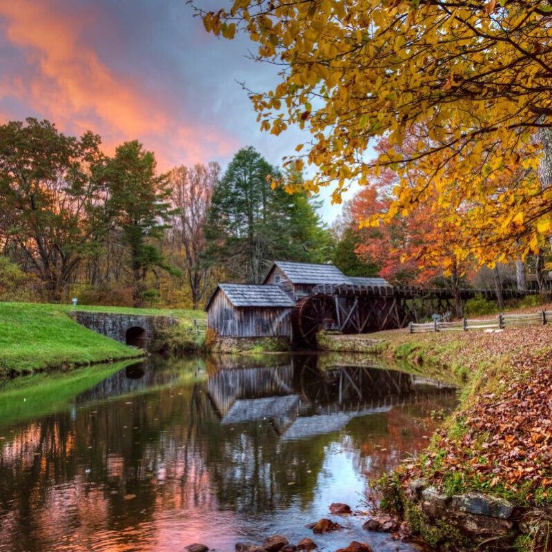 The sun sets behind the historic gristmill at Mabry Mill in Autumn, with pink highlights in the sky and red and yellow leaves framing the millpond.