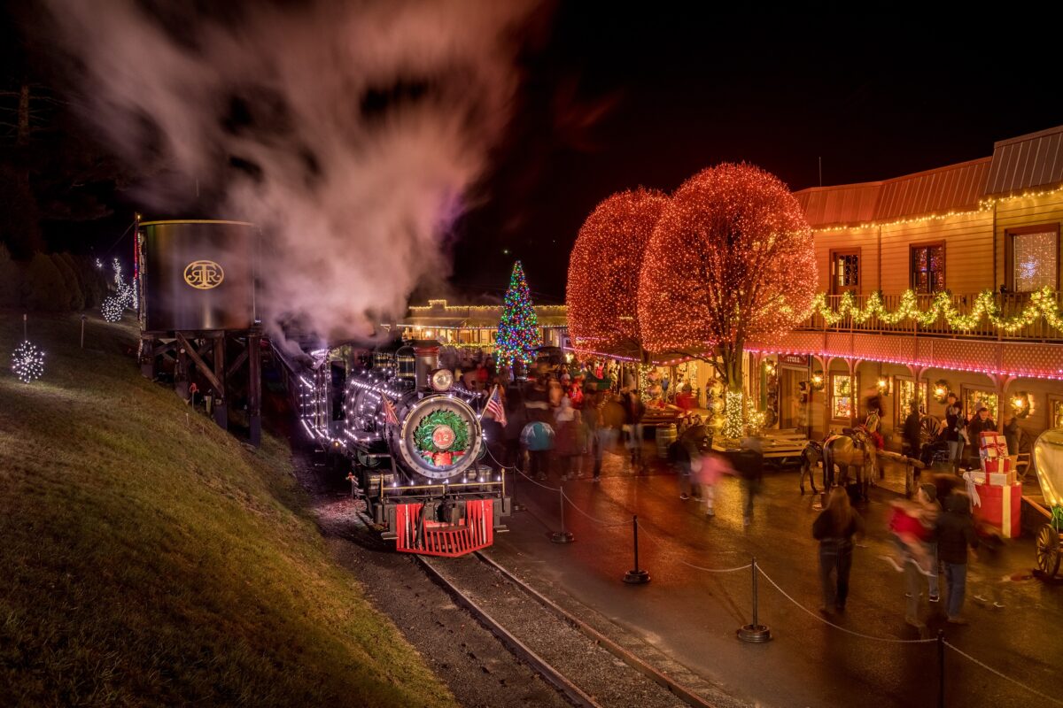 Smoke rises from the Tweetsie Train lit with Christmas lights as it sits alongside a brightly lit avenue.