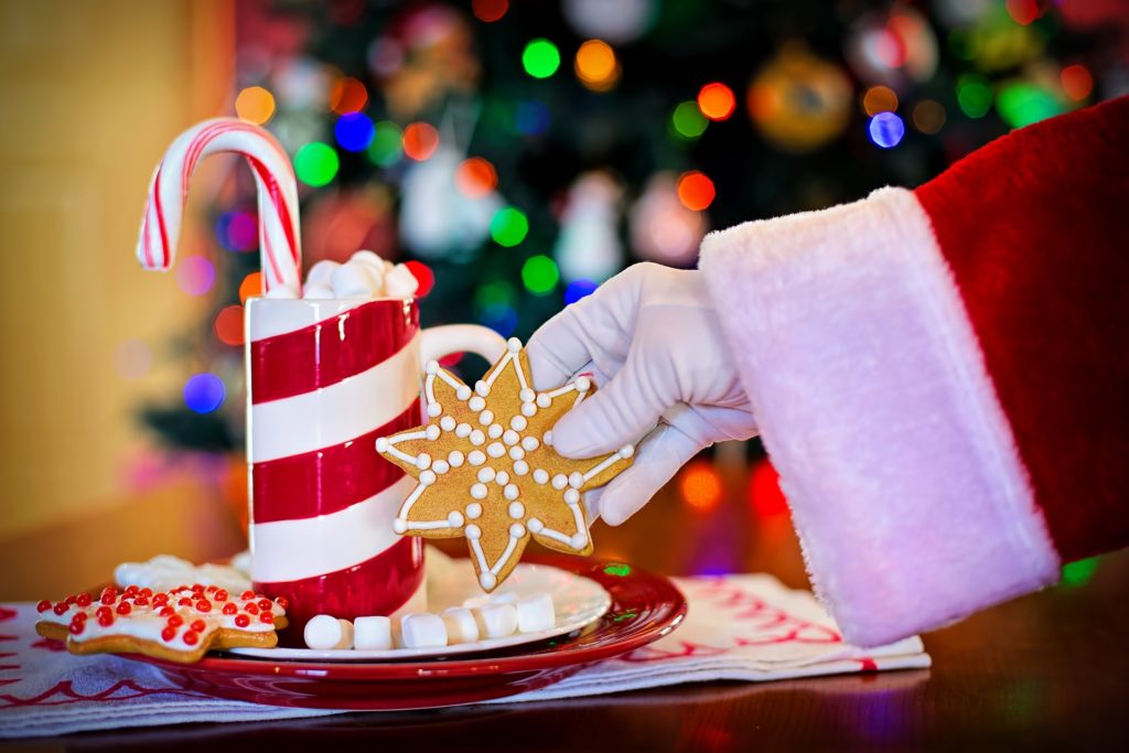 Santa's hand holds a decorated cookie next to a candy-cane striped mug topped with a candy cane and miniature marshmallows.