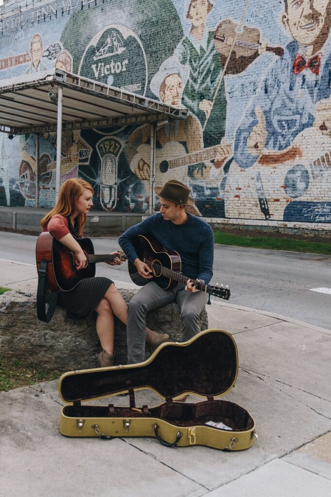 A couple sit on a rock playing guitars outdoors with an open guitar case at their feet and a mural of musicians on the building behind them.