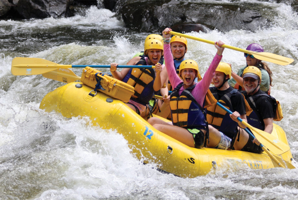 Several people wearing yellow life jackets and holding paddles sit in a river raft in a whitewater rapid.