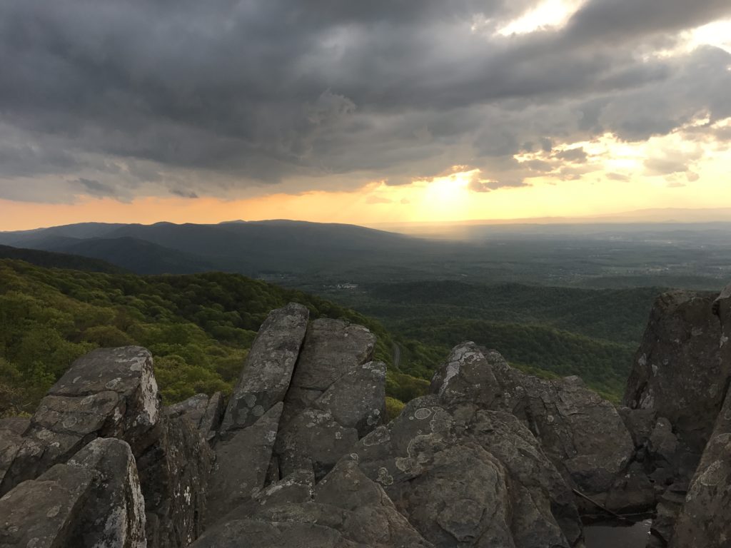 The sun sets over the valley stretching away from the rocky overlook while storm clouds loom overhead.