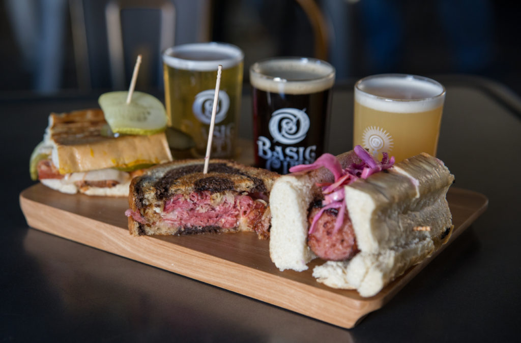 Three flavors of local beer at Hops Kitchen sit atop a sandwich board with three brewery-style sandwiches.