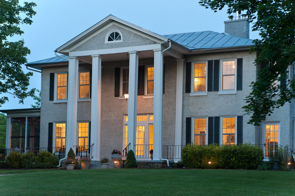 Light spills through the windows of the two story Steeles Tavern Manor front windows through the columns supporting the high porch roof.