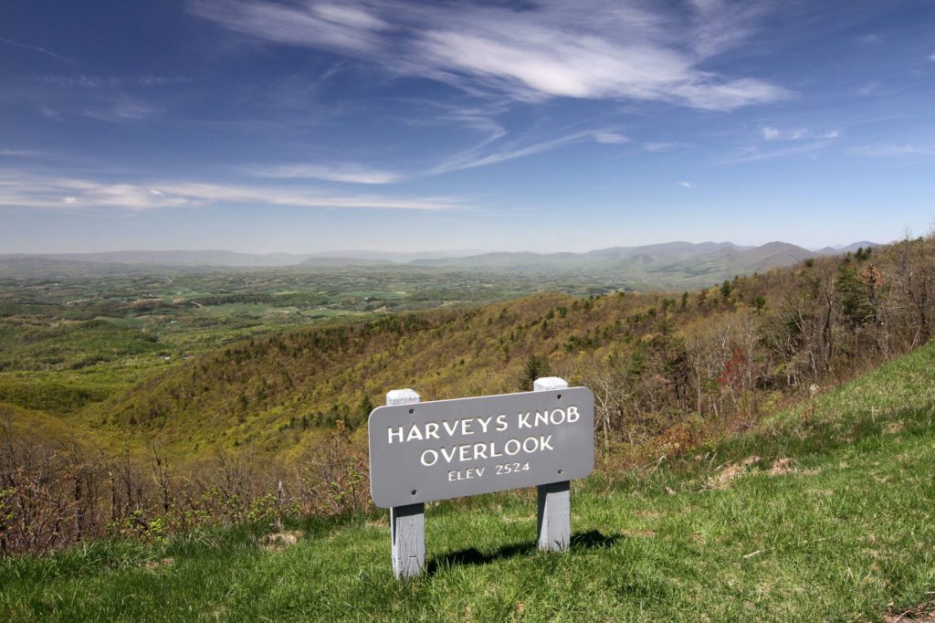 Harveys Knob Overlook reveals a sunny wide green valley alternating in forest and grasses with wispy cirrus clouds overhead.