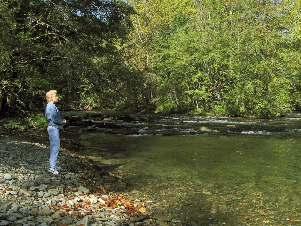 A woman stands on rounded rocks on the shore of the Oconaluftee River as it rushes over the rocky bottom.