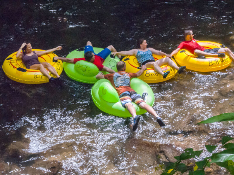 Visitors in t-shirts and shorts recline in brightly colored inner tubes in a rocky river.
