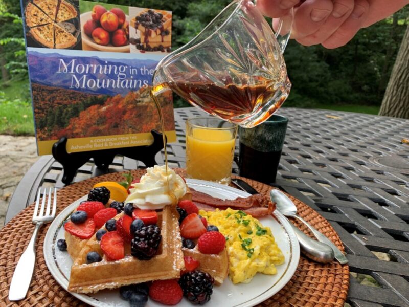 A breakfast cookbook sits behind a plate of fruit, waffles, omelet and bacon with a glass of orange juice and a pitcher of syrup.