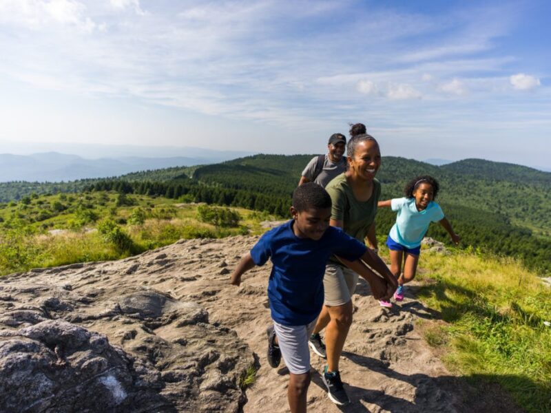 A smiling family enjoys hiking over rocky terrain with forested mountains behind them and wispy white clouds above in the cerulean blue sky.
