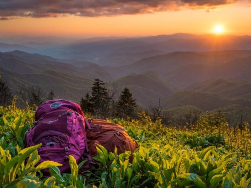 Two backpacks sit on the ground with rows of mountains framed in a golden sunset under a cloudy sky.