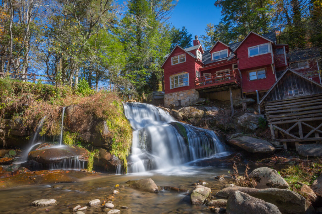 Mill Shoals Falls cascades over boulders into a serene stream with new green growth on trees above, in front of a multi-story red building.