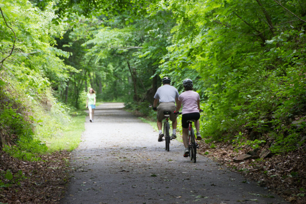 Two bikers on a tree-lined paved path in Lynchburg, VA.
