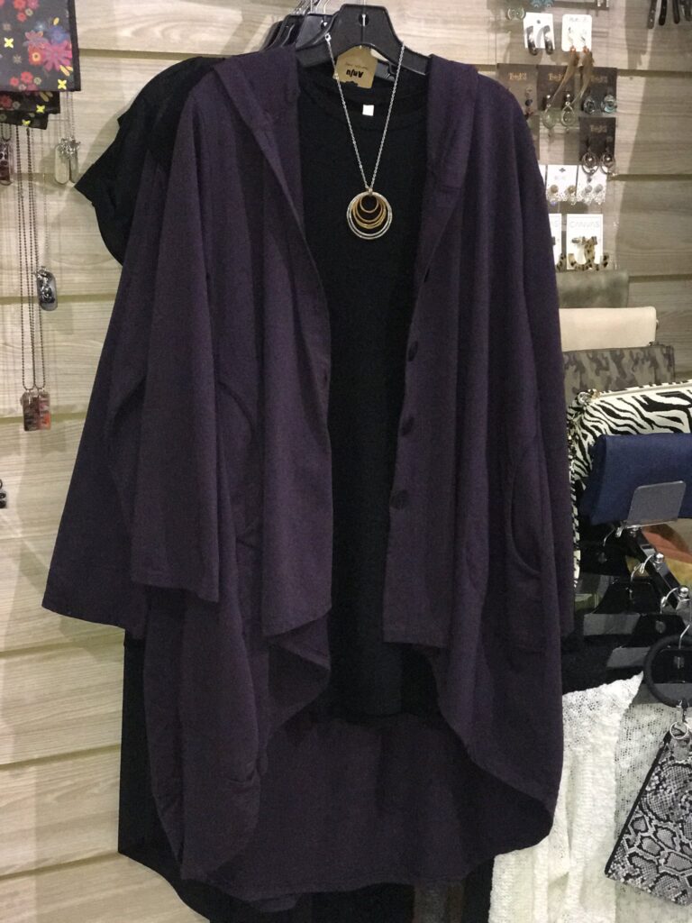 A purple women's sweater hangs over a black shirt and necklace in a store.