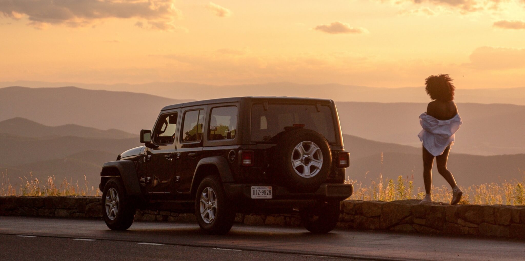 a woman walks back towards a parked black jeep as she admires a hazy golden sunset in the blue ridge parkway