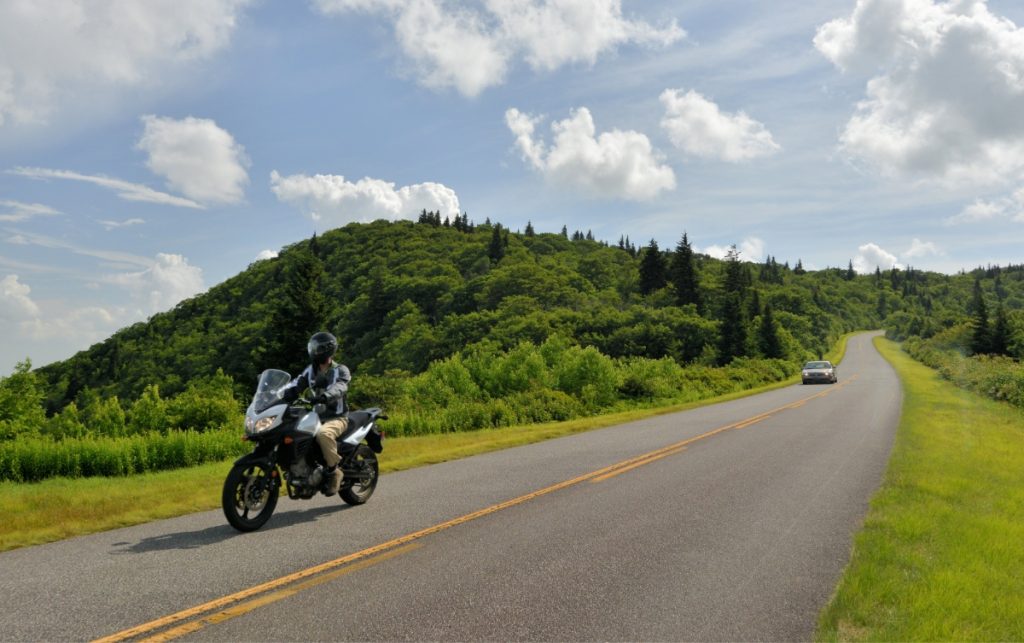 a man wearing motorcycle gear rides a black motorcycle on a paved forest road