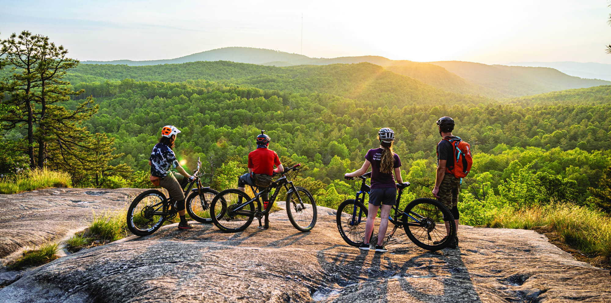 four people with bicycles admire the view of a golden sunset over the forest at a rocky viewpoint