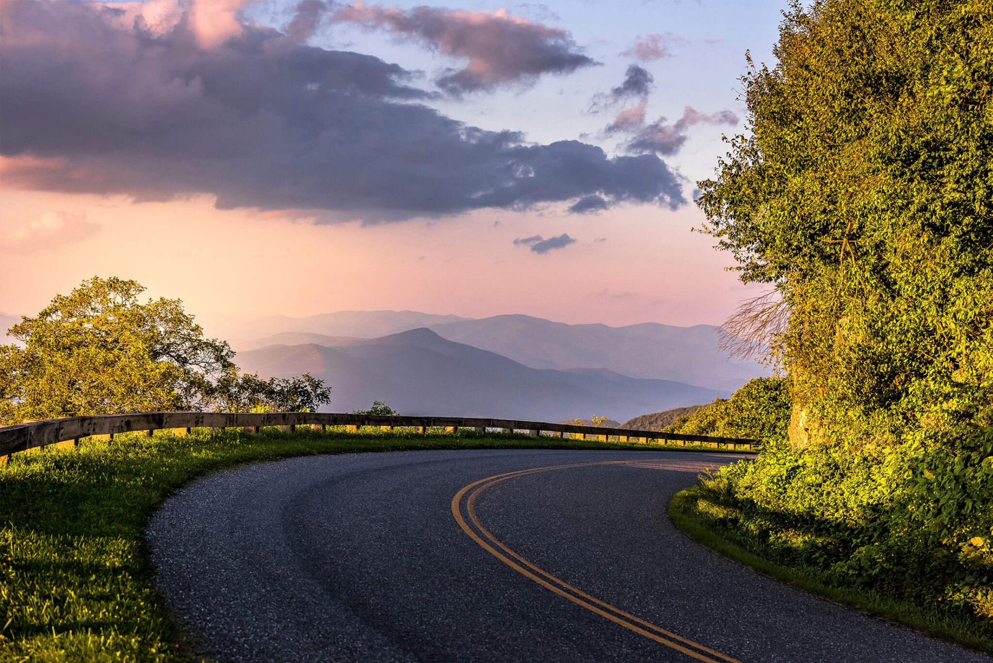 a paved road curves toward the camera with misty mountains in the background at sunset
